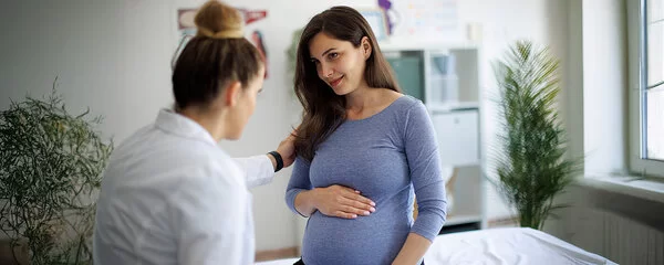 In einer Arztpraxis legt eine Frau in weißem Kittel einer Schwangeren eine Hand auf die Schulter, beide haben Blickkontakt, die Schwangere lächelt und legt eine Hand auf ihren runden Bauch.