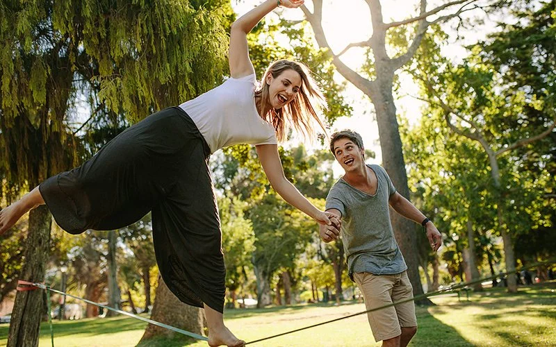 Eine Frau in lockerer Freizeitkleidung balanciert im Park auf einer Slackline. Ein Mann stützt sie mit der Hand. Beide lachen.