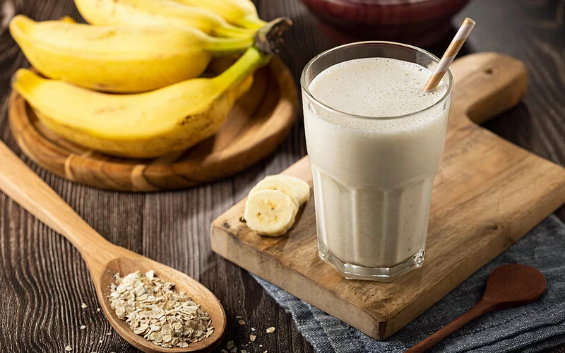 Bananen-Smoothie steht auf einem Holzbrett mit Bananenstaude und einem Löffel Haferflocken.