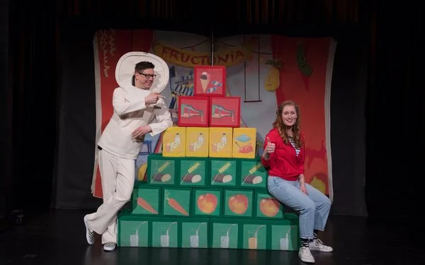 Zwei Schauspieler mit einer Ernährungspyramide in Form von Würfeln auf einer Bühne.