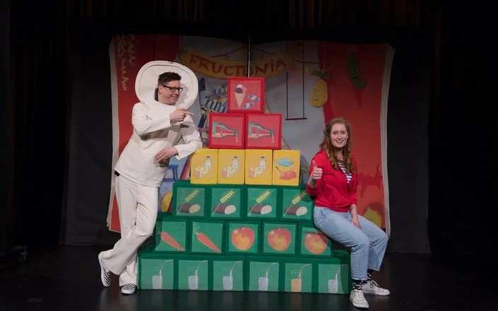 Zwei Schauspieler mit einer Ernährungspyramide in Form von Würfeln auf einer Bühne.