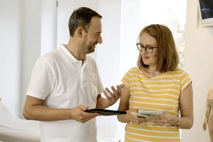 Eine Frau und ein Arzt stehen in einem Behandlungszimmer. Der Arzt hält ein Tablet in der Hand und erklärt der Frau etwas.