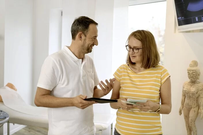 Eine Frau und ein Arzt stehen in einem Behandlungszimmer. Der Arzt hält ein Tablet in der Hand und erklärt der Frau etwas.