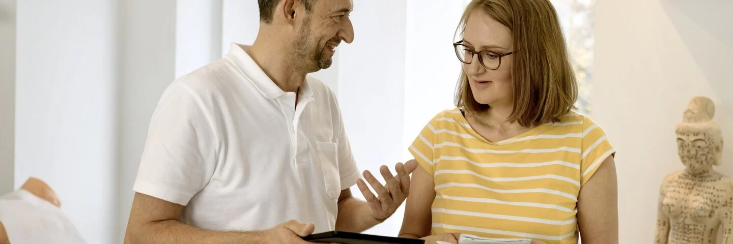 Eine Frau und ein Arzt stehen in einem Behandlungszimmer. Der Arzt hält ein Tablet in der Hand und erklärt der Frau etwas.