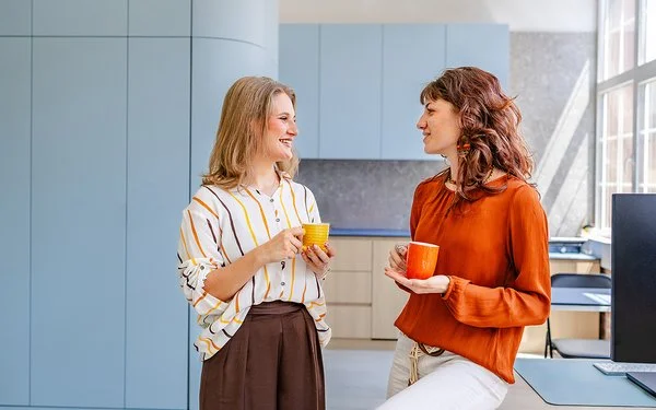 Zwei Frauen führen in einem modernen Büro ein lockeres Gespräch. Beide halten eine Kaffeetasse in der Hand.