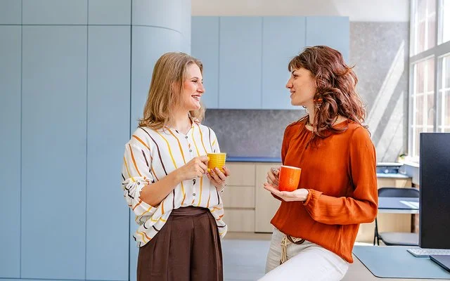 Zwei Frauen führen in einem modernen Büro ein lockeres Gespräch. Beide halten eine Kaffeetasse in der Hand.