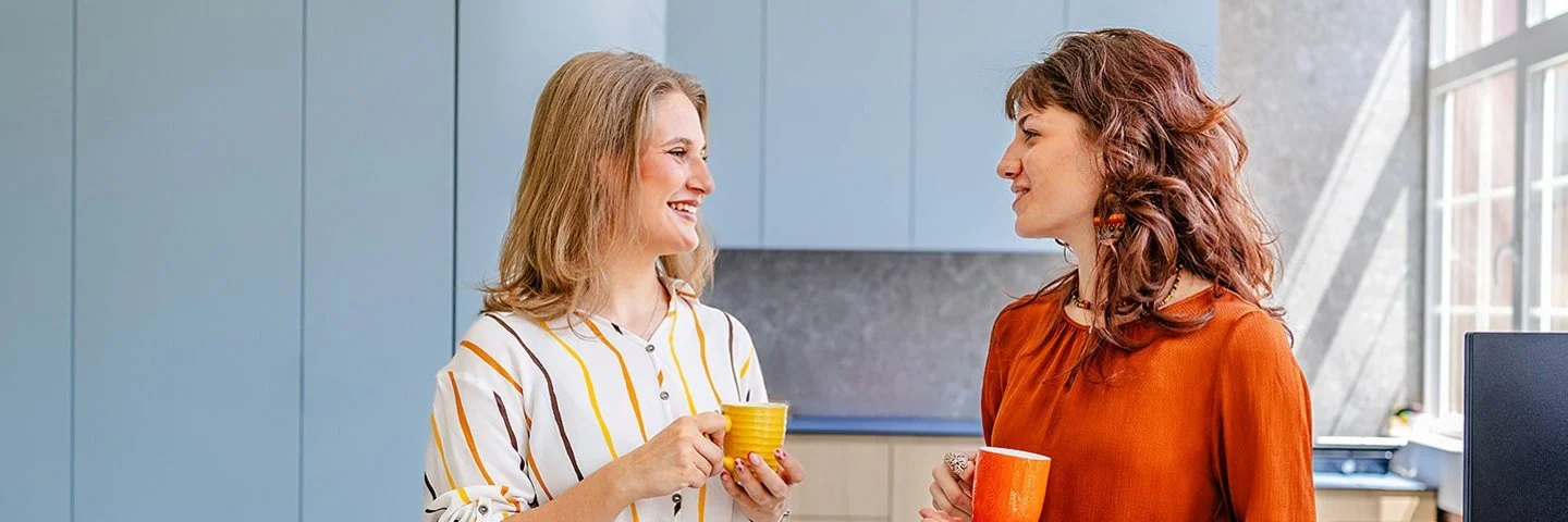 Zwei Frauen führen in einem modernen Büro ein lockeres Gespräch. Beide halten eine Kaffeetasse in der Hand.