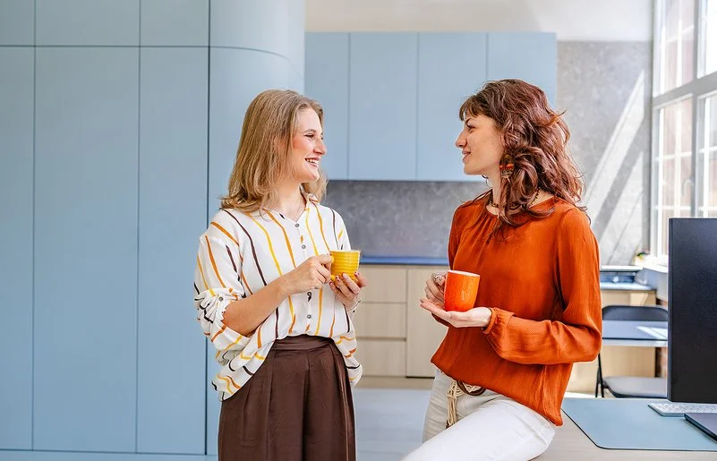 Zwei Frauen führen in einem modernen Büro ein lockeres Gespräch. Beide halten eine Kaffeetasse in der Hand.