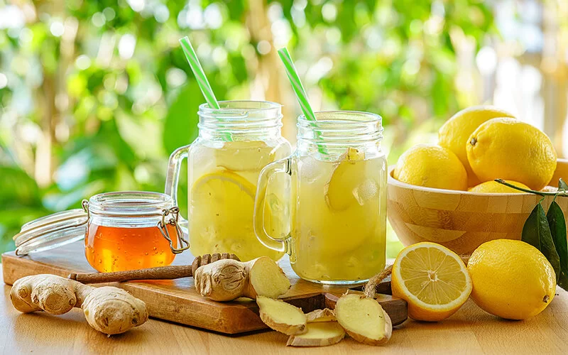 Auf einem Tisch stehen zwei Trinkgläser mit Zitronen- und Ingwerstücken darin. Daneben befinden sich Zitronen, Ingwer und ein Glas mit Honig.