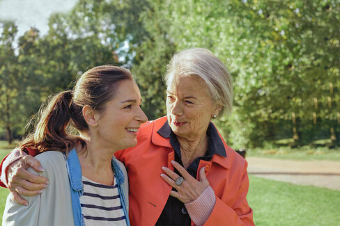 Zwei Frauen laufen durch einen Park. Die ältere Frau legt der jüngeren Frau den Arm um die Schulter und lächelt sie an.