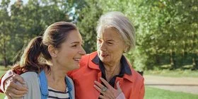 Zwei Frauen laufen durch einen Park. Die ältere Frau legt der jüngeren Frau den Arm um die Schulter und lächelt sie an.