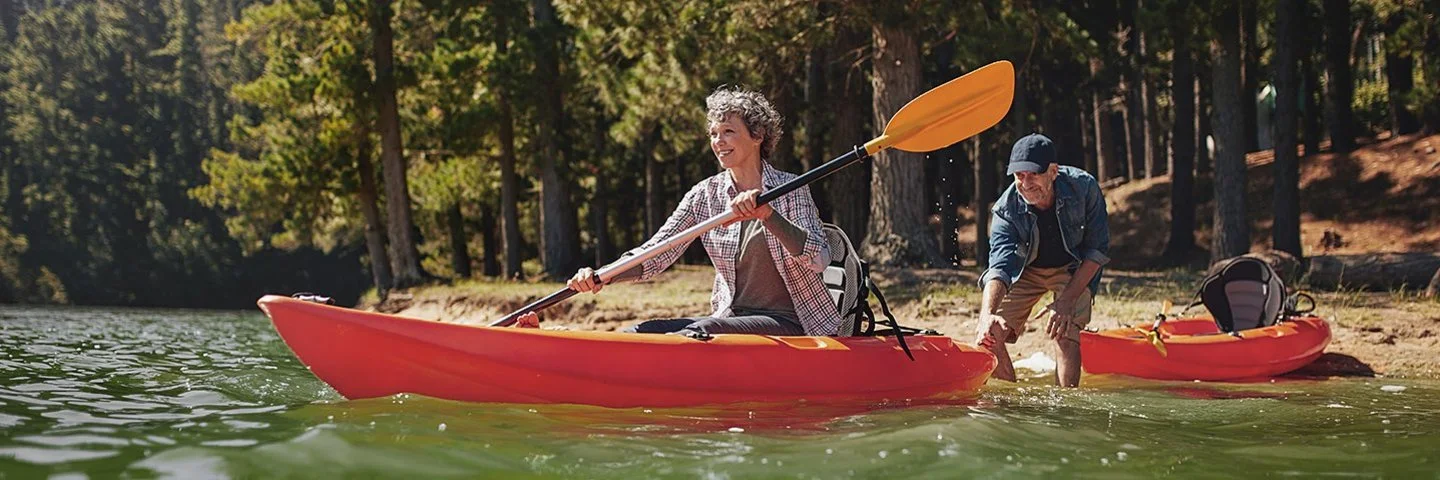 Ein Mann und eine Frau bereiten sich auf eine Kajakfahrt auf einem Fließgewässer im Wald vor. Die Frau sitzt bereits im Kajak, während der Mann sie vom Ufer aus weiter ins Wasser schiebt. Sein eigenes Kajak liegt hinter ihm am Ufer.