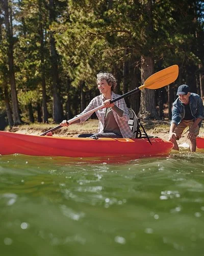 Ein Mann und eine Frau bereiten sich auf eine Kajakfahrt auf einem Fließgewässer im Wald vor. Die Frau sitzt bereits im Kajak, während der Mann sie vom Ufer aus weiter ins Wasser schiebt. Sein eigenes Kajak liegt hinter ihm am Ufer.