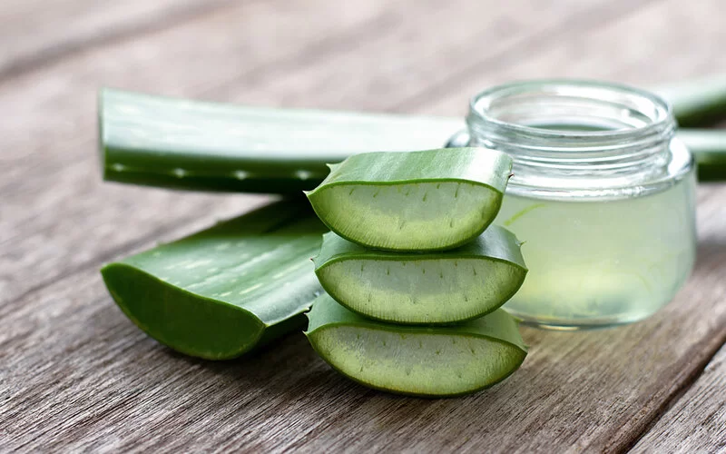 Großaufnahme von einem Holztisch, auf dem angeschnittene frische Aloe vera-Blätter liegen und ein Glas, das randvoll mit Aloe vera-Gel gefüllt ist, steht.