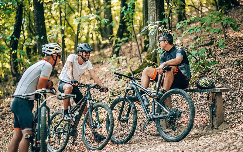Drei Männer machen im Wald auf einer Bank eine Pause vom Mountainbikefahren.