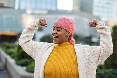 Eine lächelnde Frau mit einem rosa Kopftuch, einem gelben Rollkragenpullover und einer beigefarbenen Strickjacke macht eine Geste des Triumphes, indem sie auf beiden Seiten ihren Bizeps anspannt. Der Hintergrund ist unscharf und zeigt städtische Gebäude und Grünflächen.
