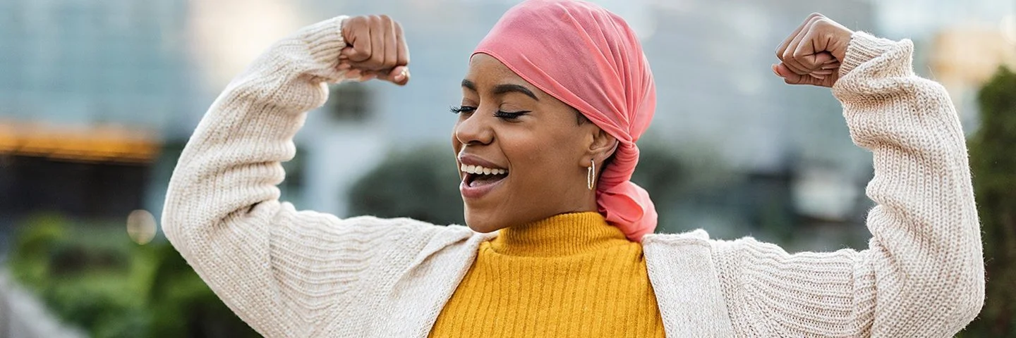 Eine lächelnde Frau mit einem rosa Kopftuch, einem gelben Rollkragenpullover und einer beigefarbenen Strickjacke macht eine Geste des Triumphes, indem sie auf beiden Seiten ihren Bizeps anspannt. Der Hintergrund ist unscharf und zeigt städtische Gebäude und Grünflächen.