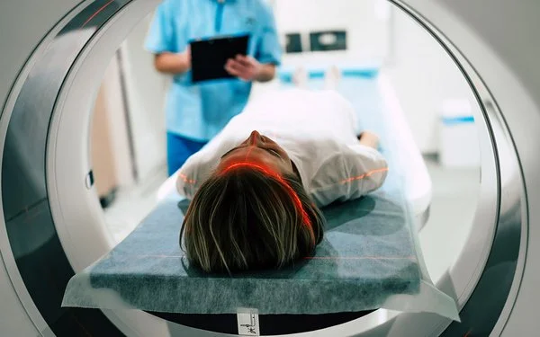 Eine junge Frau liegt vor der Öffnung eines Magnetresonanztomographen. Im Hintergrund macht jemand Notizen auf einem Papier.