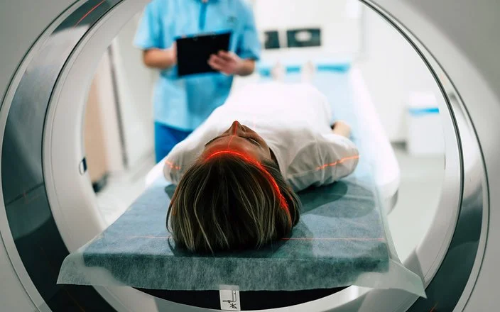 Eine junge Frau liegt vor der Öffnung eines Magnetresonanztomographen. Im Hintergrund macht jemand Notizen auf einem Papier.