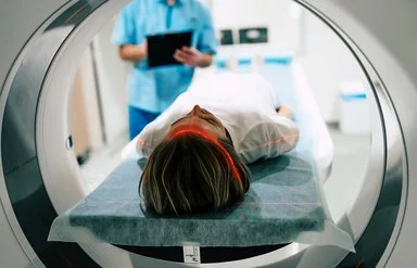 Eine junge Frau liegt vor der Öffnung eines Magnetresonanztomographen. Im Hintergrund macht jemand Notizen auf einem Papier.