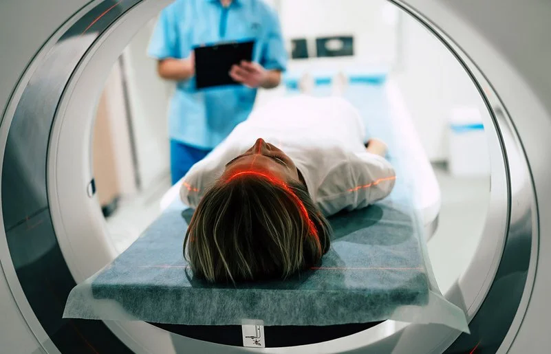 Eine junge Frau liegt vor der Öffnung eines Magnetresonanztomographen. Im Hintergrund macht jemand Notizen auf einem Papier.