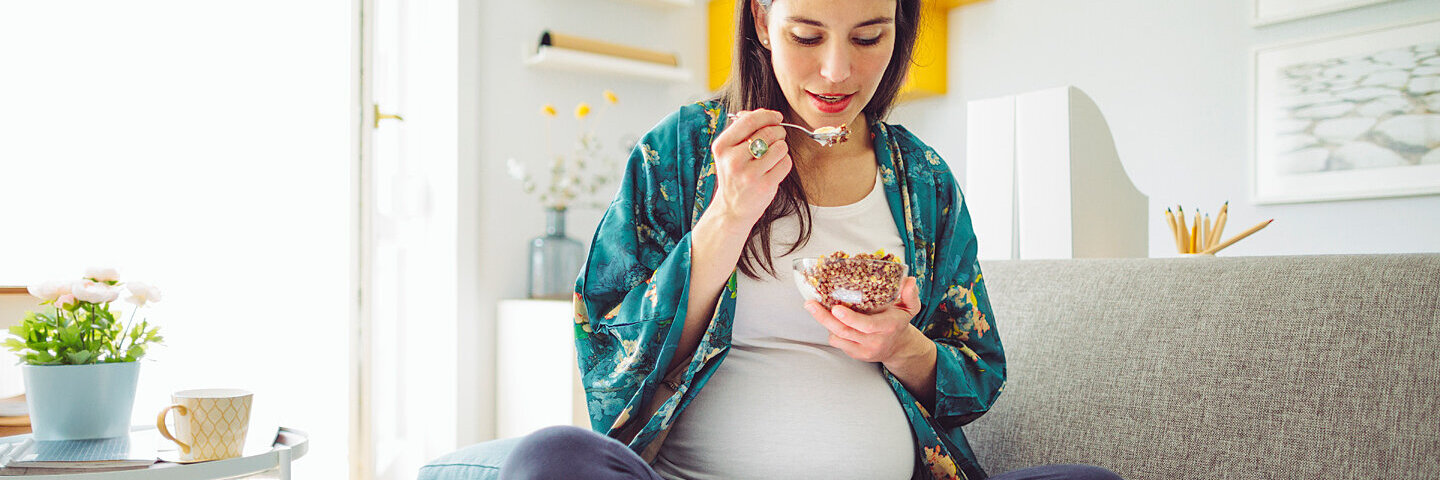 Eine schwangere Frau sitzt auf dem Sofa und isst Müsli