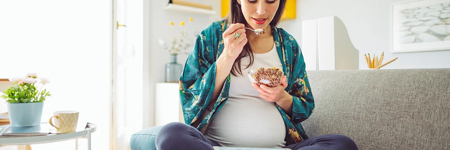 Eine schwangere Frau sitzt auf dem Sofa und isst Müsli 