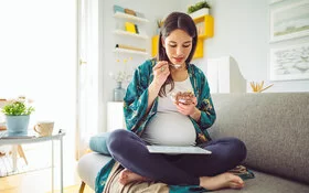 Eine schwangere Frau sitzt auf dem Sofa und isst Müsli 