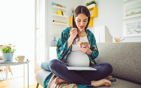 Eine schwangere Frau sitzt auf dem Sofa und isst Müsli 