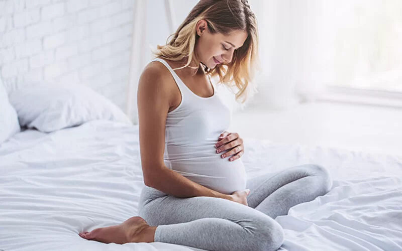 Eine Schwangere sitzt auf dem Bett und hält ihren Bauch. Sie denkt über die Coronaimpfung und Impfmythen nach.