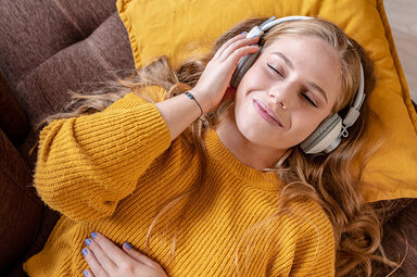Eine junge Frau mit einem gelben Pullover hört über ihre Kopfhörer Musik.