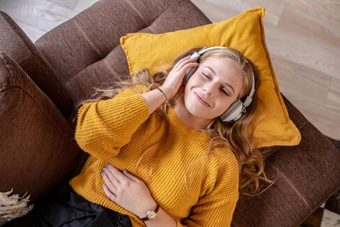 Eine junge Frau mit einem gelben Pullover hört über ihre Kopfhörer Musik. 