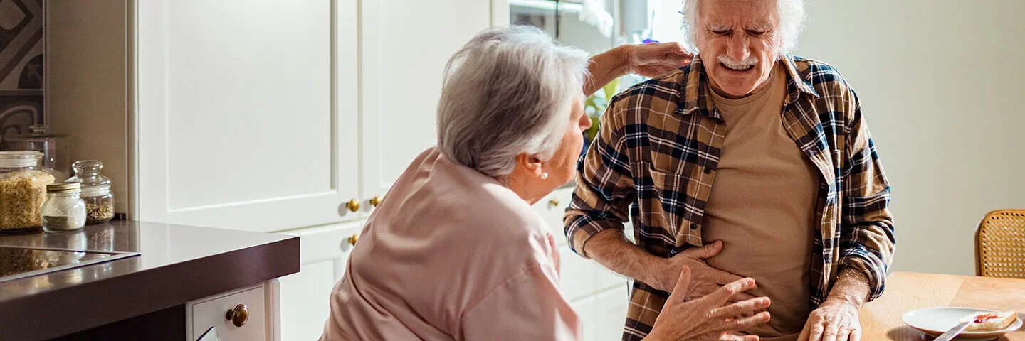 Ein älterer Mann steht mit seiner Frau in Küche und hat durch eine Divertikulitis plötzliche Schmerzen im Unterbauch.