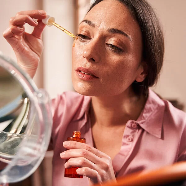 Eine Frau sitzt vor einem Kosmetikspiegel und trägt unter ihrem rechten Auge mit einer Pipette ein Serum auf.