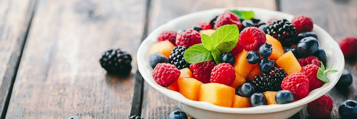 Eine Schüssel mit frischem Obst steht auf einem Holztisch, daneben liegen Beeren.