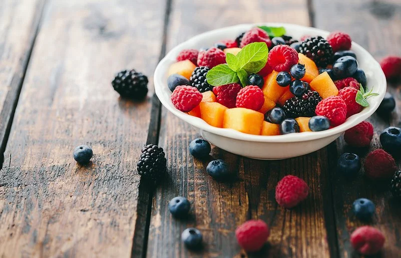 Eine Schüssel mit frischem Obst steht auf einem Holztisch, daneben liegen Beeren.