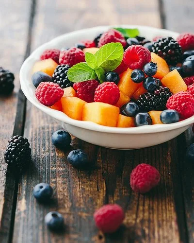 Eine Schüssel mit frischem Obst steht auf einem Holztisch, daneben liegen Beeren.