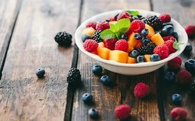 Eine Schüssel mit frischem Obst steht auf einem Holztisch, daneben liegen Beeren.