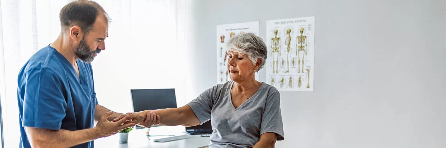 Eine Seniorin sitzt auf einer Untersuchungsliege, während ein Arzt ihre rechte Hand untersucht.