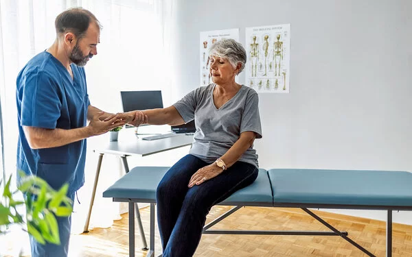 Eine Seniorin sitzt auf einer Untersuchungsliege, während ein Arzt ihre rechte Hand untersucht.