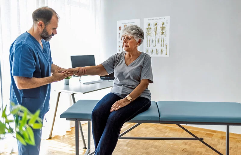 Eine Seniorin sitzt auf einer Untersuchungsliege, während ein Arzt ihre rechte Hand untersucht.
