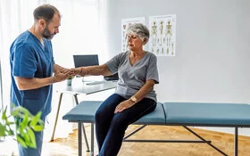 Eine Seniorin sitzt auf einer Untersuchungsliege, während ein Arzt ihre rechte Hand untersucht.