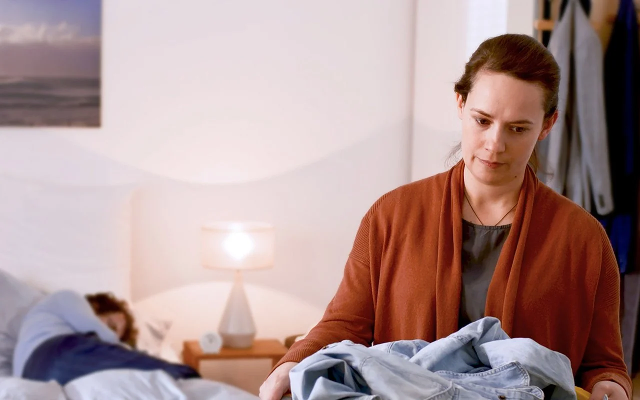 Eine Frau trägt einen Wäschekorb voller Kleidungsstücke, sie wirkt müde. Im Hintergrund ist ein im Bett liegender Mann zu erkennen.