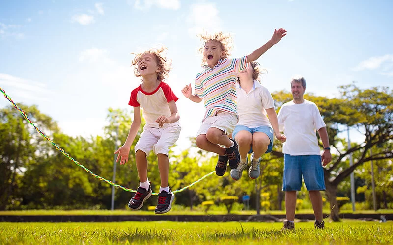 Auf einer sommerlichen Wiese springen drei Kinder gemeinsam über ein Seil. Im Hintergrund steht ein Mann, der das Seil auf einer Seite festhält.