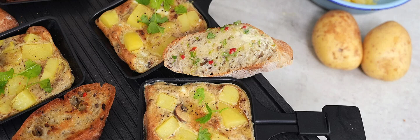 Pfännchen-Frittata und vegane Kräuterbutter mit Baguette nach einem Rezept von Felicitas Then, angerichtet auf einem Raclettegrill. Rechts daneben eine hellblaue Schale mit Kartoffelscheiben, davor zwei Kartoffeln.