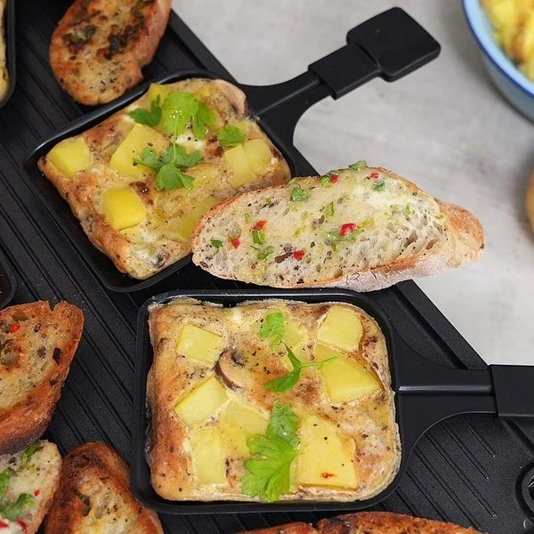 Pfännchen-Frittata und vegane Kräuterbutter mit Baguette nach einem Rezept von Felicitas Then, angerichtet auf einem Raclettegrill. Rechts daneben eine hellblaue Schale mit Kartoffelscheiben, davor zwei Kartoffeln.