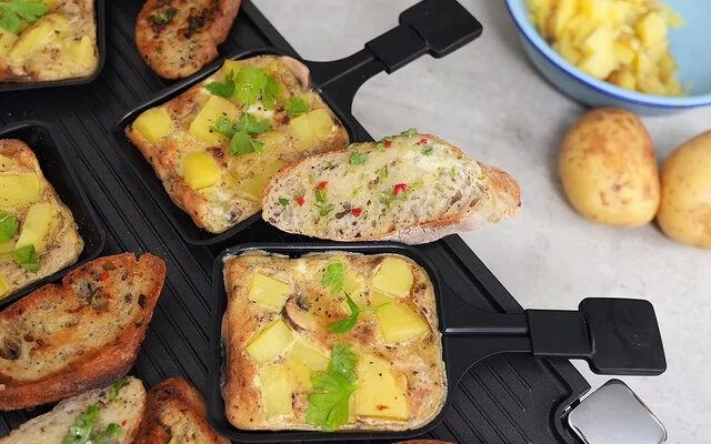 Pfännchen-Frittata und vegane Kräuterbutter mit Baguette nach einem Rezept von Felicitas Then, angerichtet auf einem Raclettegrill. Rechts daneben eine hellblaue Schale mit Kartoffelscheiben, davor zwei Kartoffeln.