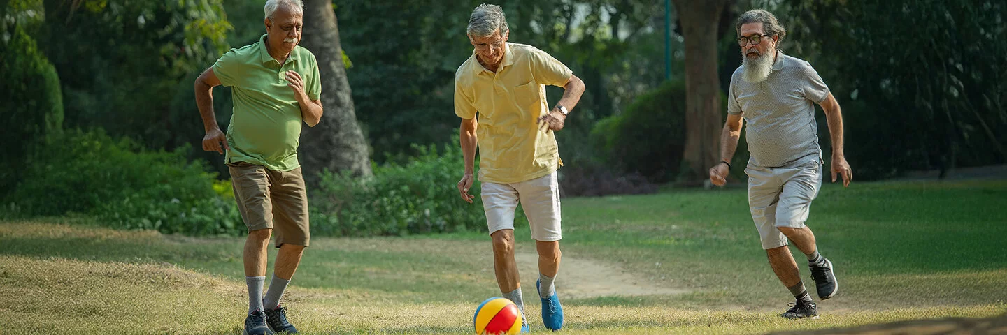 Drei ältere Männer spielen auf einer Wiese Walking Football, der mittlere Mann schießt gerade den Ball.