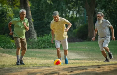 Drei ältere Männer spielen auf einer Wiese Walking Football, der mittlere Mann schießt gerade den Ball.