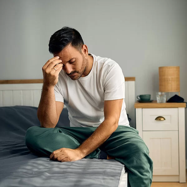 Ein junger Mann sitzt auf seinem Bett im Schlafzimmer. Mit gesenktem Kopf stützt er den rechten Ellenbogen auf dem rechten Knie ab und fasst sich mit der rechten Hand an die Stirn.